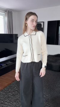 Cardigan blanc en tricot avec bordure noire, deux poches et un col ; associé à un pantalon large noir, dans un intérieur minimaliste.