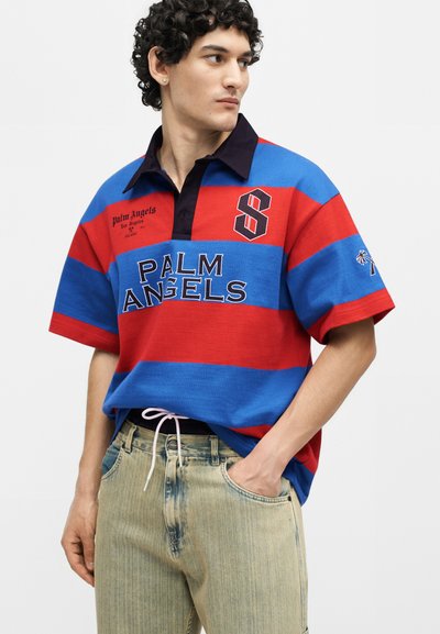 Νέος άνδρας που φοράει μπλε και κόκκινο ριγέ πόλο με το κείμενο "Palm Angels", ανοιχτόχρωμο τζιν και ένα χέρι στην τσέπη, κοιτάζει προς τα δεξιά.