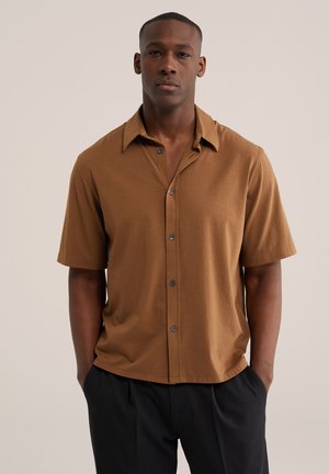 Mężczyzna ubrany w brązową, krótkorękawową koszulę na guziki i czarne spodnie, stojący z rękami w kieszeniach na jednolitym jasnym tle.