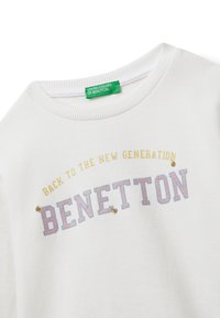 Sudadera blanca con escote redondeado, con texto multicolor "VUELTA A LA NUEVA GENERACIÓN" encima de "BENETTON". Etiqueta verde visible.