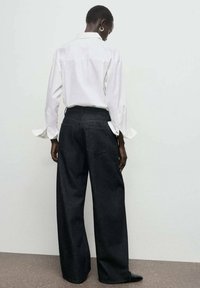 Jeans de tiro alto, de mezclilla oscura y pierna ancha, mostrados por la parte de atrás, combinados con una camisa blanca de botones con mangas largas y puños.