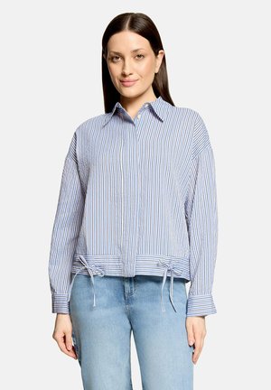 Kvinde med glat brunt hår har en blå og hvidstribet skjorte med knapper og bindebånd forneden og lyseblå jeans, stående med fronten fremad.