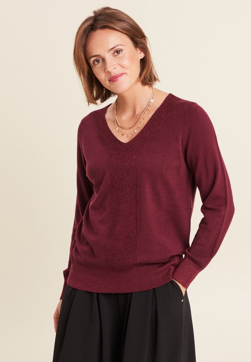 Bordeaux gebreide trui met een V-hals, voorzien van een oftextuur op het centrale paneel en lange mouwen. Gecombineerd met een zwarte rok, die de vorm accentueert.