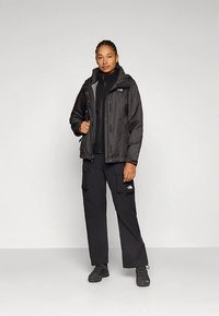 Must veekindel jope kapuutsiga, esikinnitus lukuga ja küljetaskutega, koos mustade cargo-pükstega. Tugev materjal, mõeldud välitingimustes kasutamiseks.