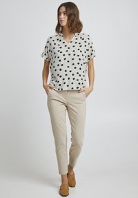 Blusa a maniche corte, scollo a V in beige chiaro con pois neri, abbinata a pantaloni slim beige e mocassini marroni con dettagli in metallo dorato.