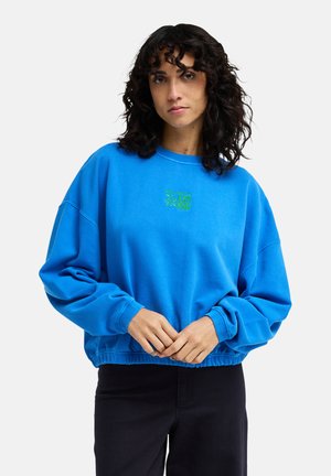 Vrouw met krullend donker haar, een felblauwe oversized sweatshirt met een groen logo, handen voor zich samengevouwen, staand tegen een effen achtergrond.
