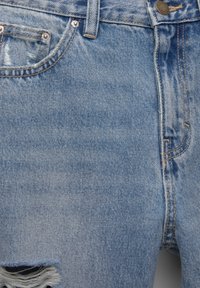 Jean en denim bleu clair en gros plan montrant la poche avant, la passants de ceinture, le bouton et une déchirure effilochée près du bord inférieur gauche.
