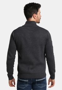 Dunkelgrauer gestrickter Pullover mit hohem Kragen, gerippten Bündchen und Saum. Verfügt über ein Patch an den linken Ärmel. Getragen mit blauen Denim-Jeans.