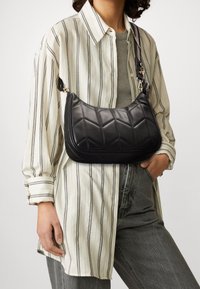 Quilted sort læder skuldertaske med en afrundet form, guldfarvede hardware-detaljer og en aftagelig strop, båret over en stribet skjorte.