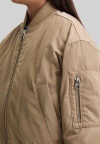 Beige Bomberjacke mit geripptem Kragen, Frontreißverschluss und seitlichem Reißverschlussfach. Glatte Textur und gepolstertes Design für Wärme.