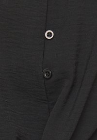 Camicia in tessuto nero con superficie testurizzata, dotata di due bottoni neri e dettagli in bianco. Il design comprende strati di tessuto sovrapposti.