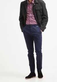 Donker jasje met twee borstzakken, rood-wit geruite blouse, marineblauwe slim-fit broek en zwart suède schoenen met lichte zolen.