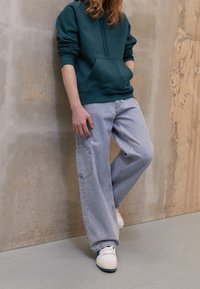 Dunkelgrüne Kapuzenjacke, lockere hellblaue Jeans mit Seitentasche und weiße Sneakers, die an einer strukturierten Betonwand lehnen.