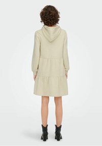 Vestido con capucha y mangas largas en tela beige clara, con falda de capas. Diseño sencillo; no hay patrones ni acentos visibles. Se llevan botas negras hasta los tobillos.