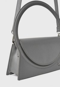 Borsa grigia con una base triangolare, manico circolare in cima, superficie texturizzata e dettagli metallici. Presenta una chiusura a scomparsa e una tracolla regolabile.