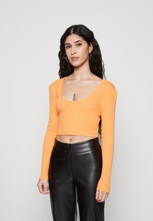 Oransje langermet crop topp med bred halsåpning, kombinert med høytlivede svarte skinnbukser. Glatt tekstur, tettsittende design.