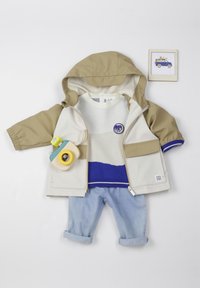 Giacca cappuccio beige, maglione bianco con striscia blu, jeans azzurri arrotolati e un giocattolo macchina fotografica in legno. Layout chiaro e strutturato.