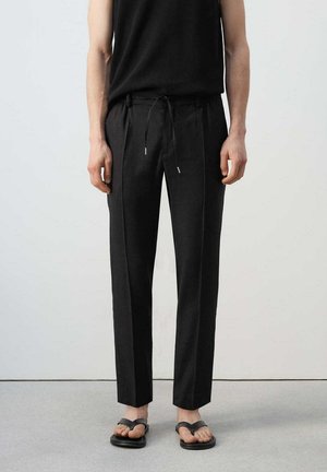 Osoba ubrana w czarne spodnie ze sznurkiem, czarny top bez rękawów oraz czarne klapki, stojąca na tle jasnego, jednolitego tła.