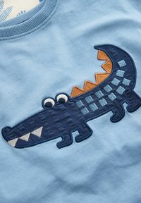 Lichtblauwe sweatshirts met een borduursel van een krokodil in donkerblauw met oranje accenten, getextureerde schubben en grote witte ogen.