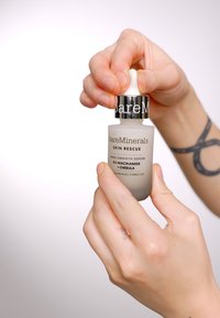 Siero Skin Rescue di BareMinerals in una bottiglia di vetro frosted, bianca con testo nero, tappo contagocce argento. Mano che tiene il contagocce.