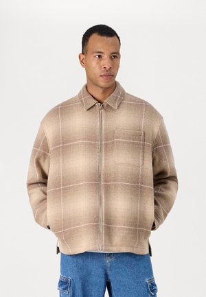Beige plaid jacket with a zip front, collar, and chest pocket. Made from textured fabric, features white grid lines and a relaxed fit.