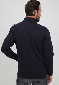 Felpa blu navy con colletto alto, maniche lunghe e tessuto strutturato; presenta un dettaglio di cucitura curva sul retro.