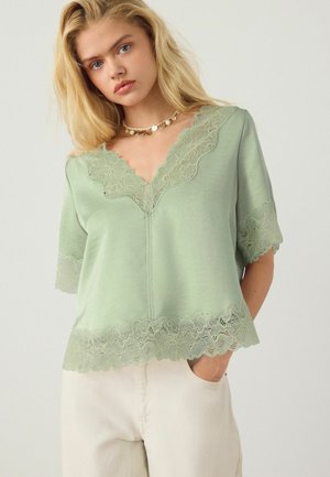 Szőke nő világoszöld, csipkés V-nyakú felsőben és fehér nadrágban, arany pénzérme nyaklánccal, zsebre tett kézzel, egyszínű háttér előtt.