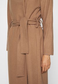 Primo piano di una persona che indossa un cappotto di lana marrone con una cintura annodata in vita e maniche lunghe.