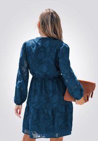 Abito blu scuro a strati con sovrapposizione floreale trasparente, maniche lunghe, vita elastica e scollo rotondo. Tiene una borsa a pochette marrone.