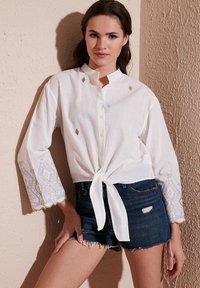 Chemise blanche à boutons avec un nœud à l'avant, présentant des accents brodés en bleu et or sur les manches évasées, associée à un short en jean déchiré.