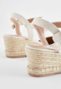 Sandales espadrilles compensées avec talons en jute tissé et brides de cheville en tissu beige sur fond blanc.