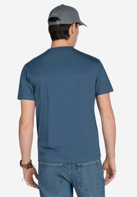 Camiseta de manga corta azul hecha de tela suave, con cuello redondo y diseño liso. Usada con jeans de mezclilla azul claro y una gorra gris.