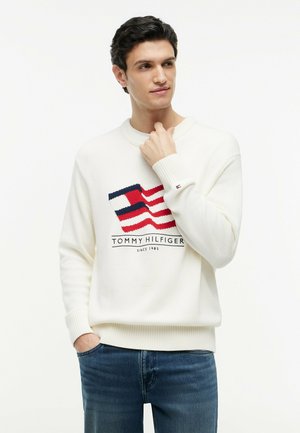 Joven con un suéter blanco de Tommy Hilfiger con un logo rojo, blanco y azul y vaqueros azules, mirando hacia un lado.