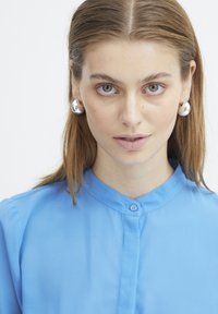 Camicia azzurra con colletto, caratterizzata da una texture liscia. Il modello indossa orecchini tondi in argento lucido. Sfondo chiaro.