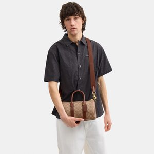Persona que lleva una camisa negra de manga corta con estampado y pantalones blancos, sosteniendo un bolso marrón con monograma y asa para el hombro.