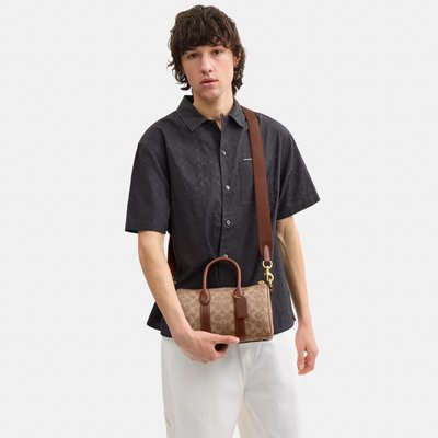 Personne portant une chemise noire à manches courtes avec un motif et un pantalon blanc, tenant un sac à main marron monogrammé avec une bandoulière.