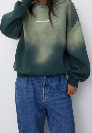 Person, die einen grün-blauen Batik-Sweatshirt mit dem Schriftzug „nostalgische Stimmung“ trägt, kombiniert mit lockeren blauen Denim-Jeans vor weißem Hintergrund.