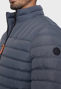 Giacca imbottita di colore grigio scuro con sezioni a trama orizzontale, colletto alto e chiusura con zip, con dettaglio arancione e patch del logo nero.