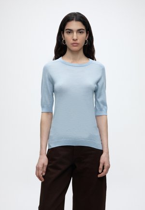 Femme aux longs cheveux foncés portant des boucles d'oreilles créoles dorées, un pull léger bleu clair à manches courtes et un pantalon marron foncé sur un fond uni.