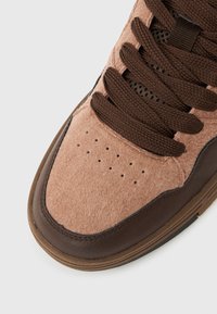 Κοντινό πλάνο μύτης αθλητικού παπουτσιού από μπεζ τσόχα και σκούρο καφέ δέρμα με καφετί κορδόνια που ταιριάζουν, σε λευκό φόντο.