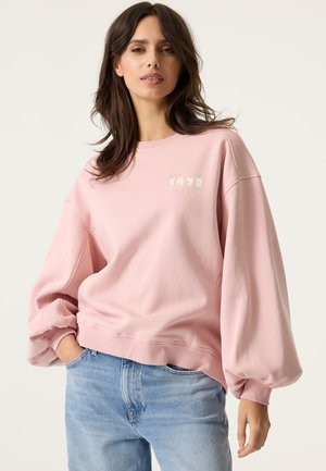 Frau mit langen dunklen Haaren, die einen rosa Pullover mit Ballonärmeln und hellblaue Jeans trägt, steht mit einer Hand in der Hosentasche.