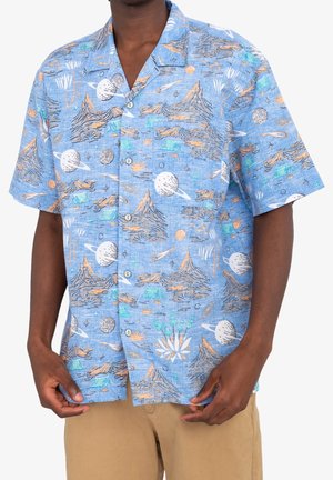 Mann trägt blaues kurzärmeliges Hemd mit Knöpfen, bedruckt mit Mustern von Planeten, Bergen und Sternen, kombiniert mit hellbraunen Hosen, steht vor einem weißen Hintergrund.