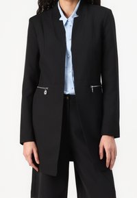 Svart kavaj med en strukturerad design, med silverfärgad dragkedjor på fickorna, skräddarsydd passform och slät textur. Kombinerad med en ljusblå skjorta under.