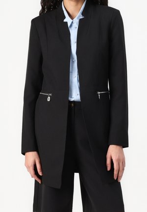 Blazer noir au design structuré, avec des fermetures éclair en argent sur les poches, coupe ajustée et texture lisse. Associé à une chemise bleu clair en dessous.