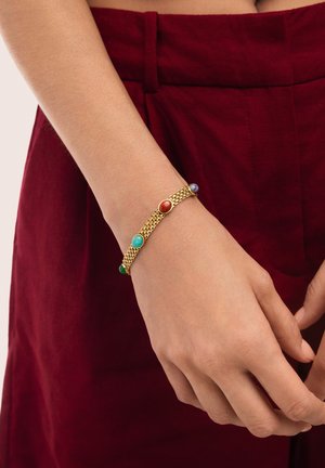 Primer plano de una mano que lleva una pulsera de cadena de oro con gemas redondas y coloridas sobre pantalones burdeos.