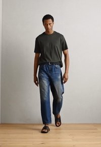T-shirt verde scuro a maniche corte, jeans in denim blu a taglio rilassato con un motivo leggermente sbiadito, e sandali aperti marroni, su uno sfondo neutro.