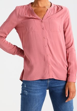 Langarm stofroze blouse met knopen en twee borstzakken, gedragen met blauwe distressed jeans, model poseert met hand op heup.