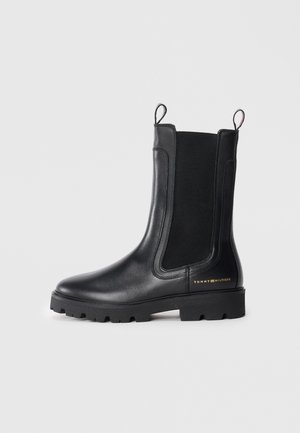 Botas de tobillo negras hechas de cuero brillante con paneles laterales elásticos, suelas de goma gruesa y tira en la parte trasera. Presenta un logotipo dorado en el lateral.
