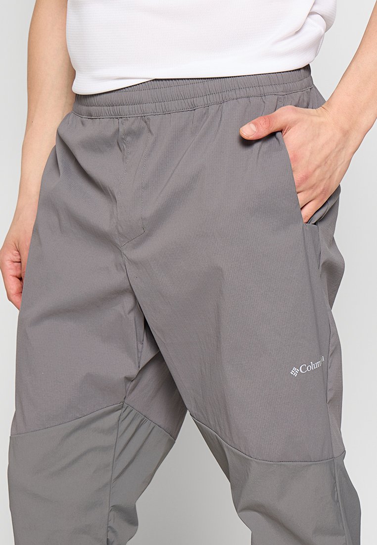 Mężczyzna w szarych spodniach Columbii z elastycznym pasem, wkładający prawą rękę do kieszeni, oraz z białą koszulą wpuszczoną w spodnie.