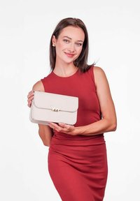 Béžová kožená kabelka se zlatým emblémem, obdélníkového tvaru a klopovým zavíráním. Modelka ji drží, oblečena v přiléhavých červených šatech. Minimalistický design.